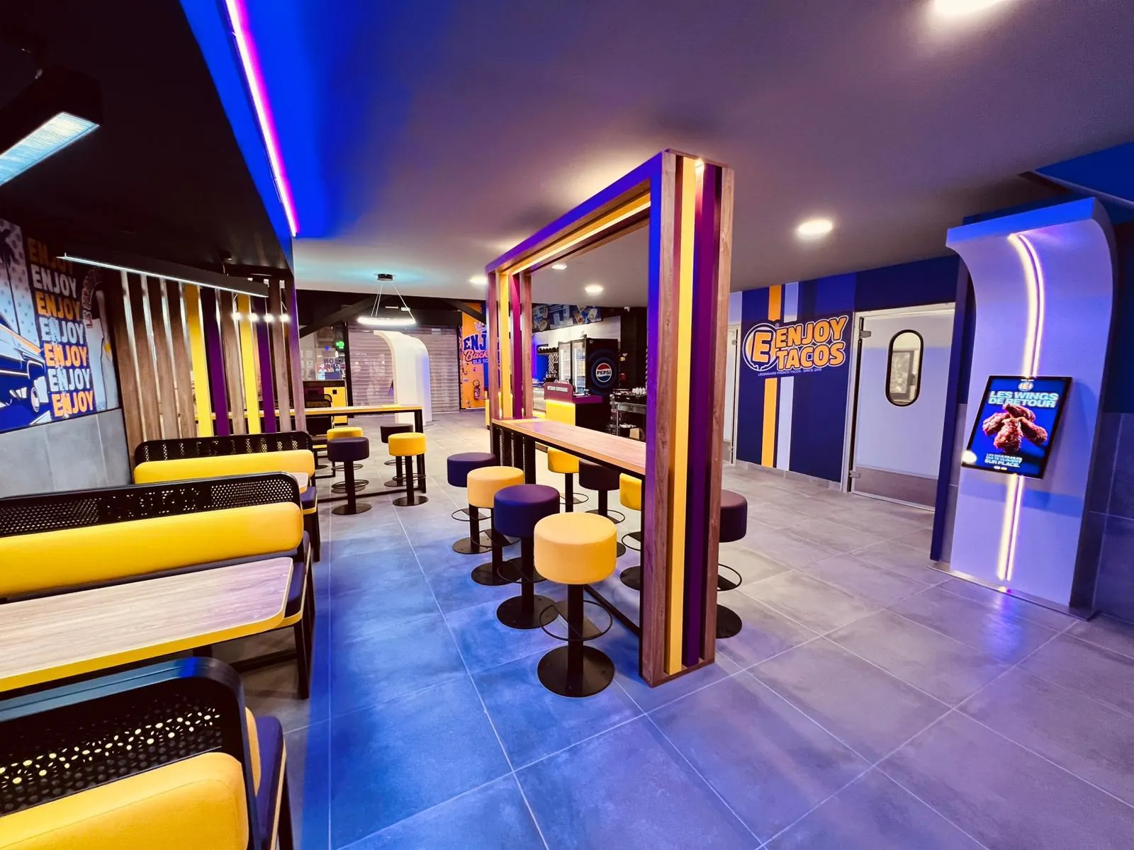 Salle d’un restaurant Enjoy Tacos : banquettes jaunes, tabourets violets et jaunes, fresque murale et néons bleus